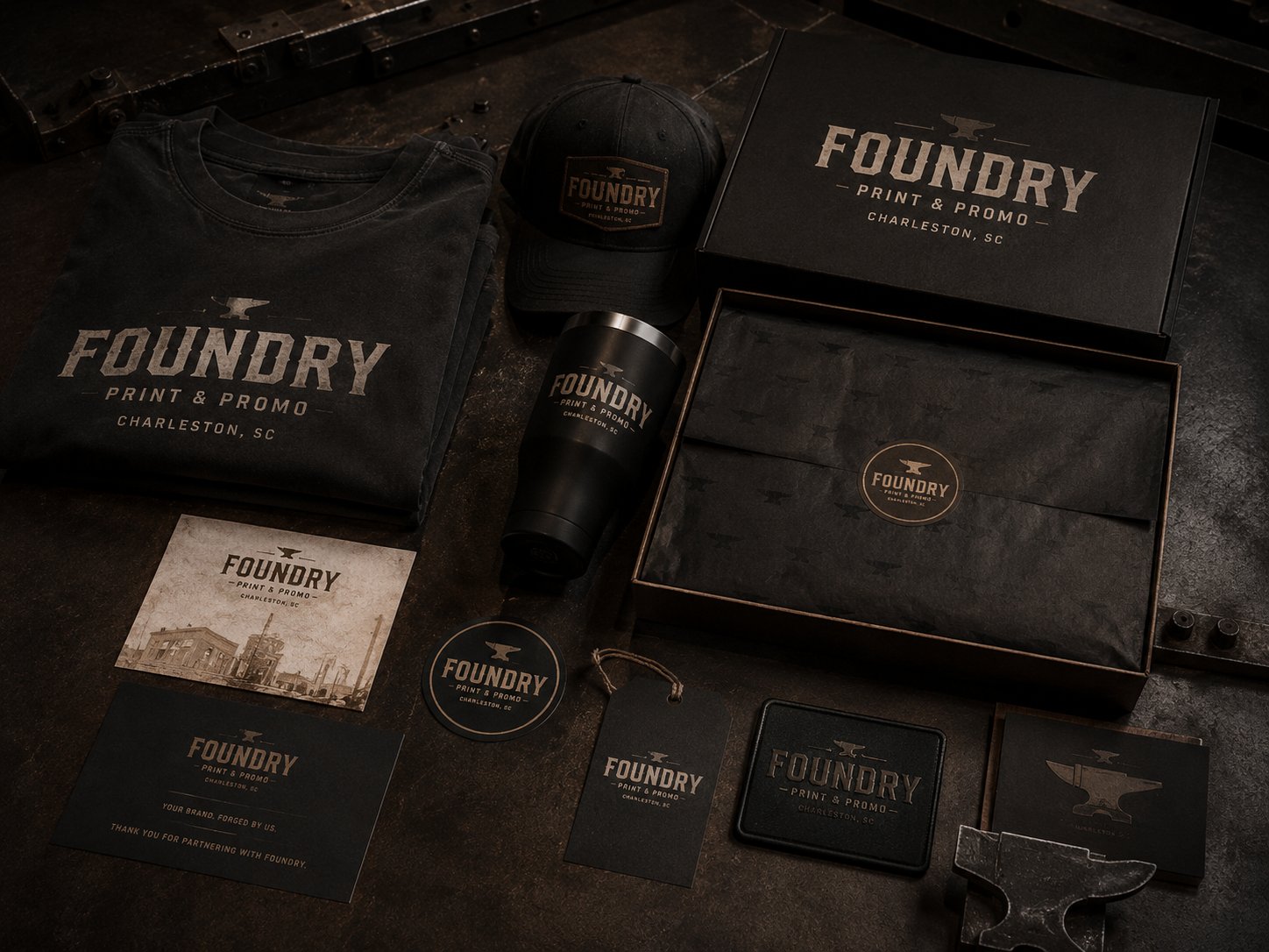 Foundry Print and Promo branded goods flat lay — shirt, hat, drinkware, packaging, and printed collateral on a dark surface